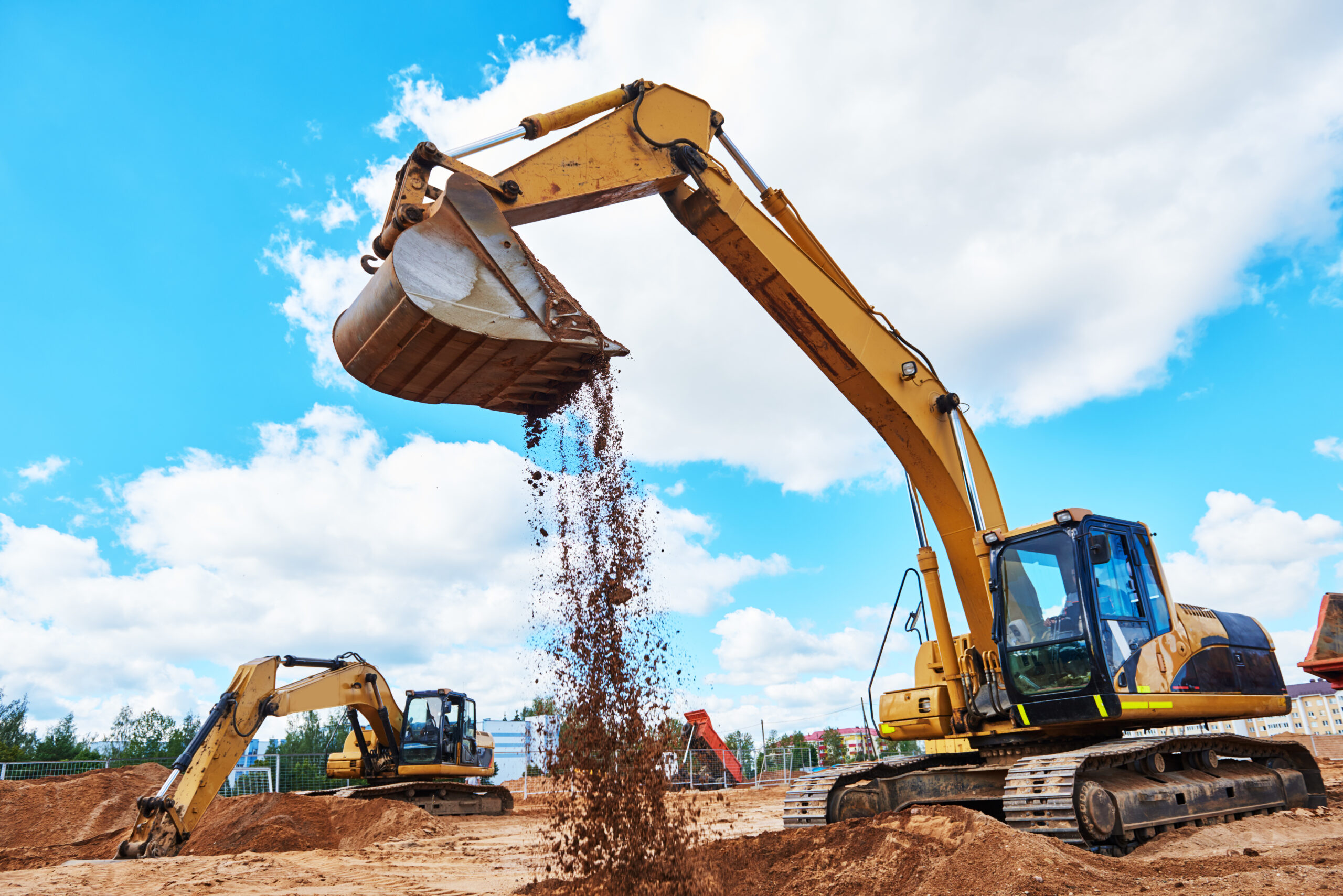 Two excavators working on a civil construction site, with one machine dumping soil from its bucket under a blue sky.