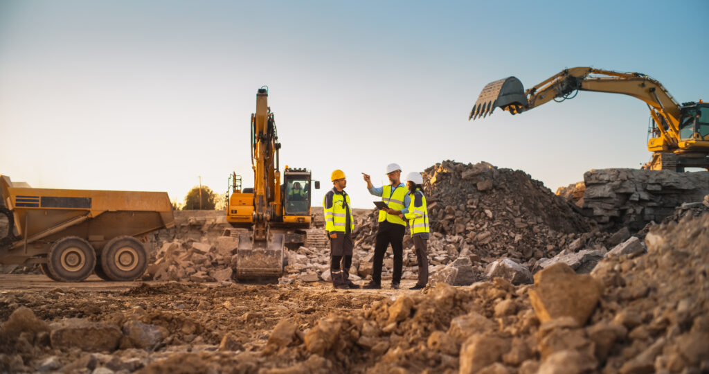 Construction managers wearing safety vests and hard hats reviewing plans on a civil construction site with excavators and a dump truck in the background.