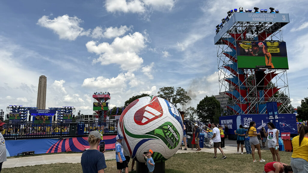 Visitors gather around a giant soccer ball at the FIFA Fan Festival in Kansas City, with event screens and the Liberty Memorial nearby.